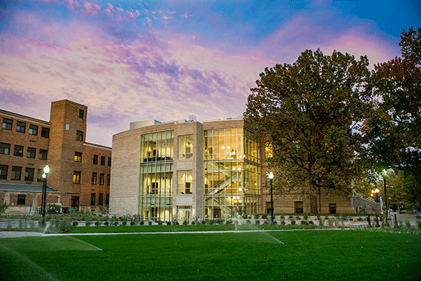 The Fine Arts Building at Indiana State is illuminated at a distance through its many glass windows beneath a pink and purple evening sky.